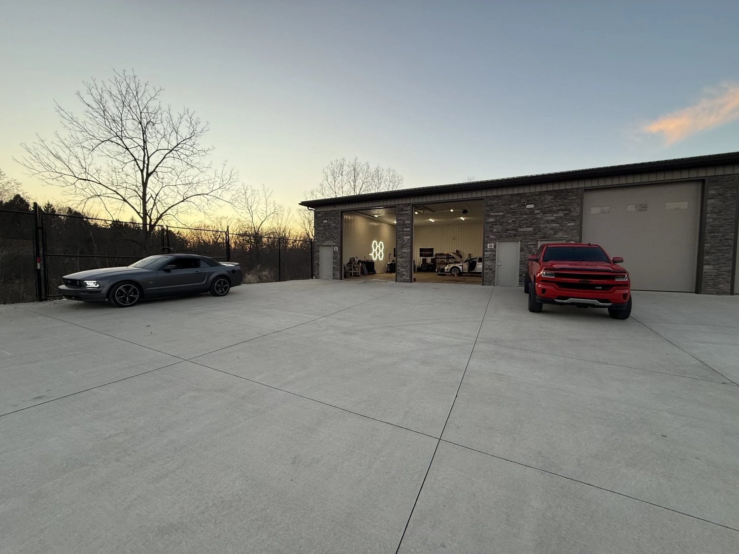 The 1Stop Auto Performance shop at dusk, bays open, with a Mustang GT and a red Silverado outside